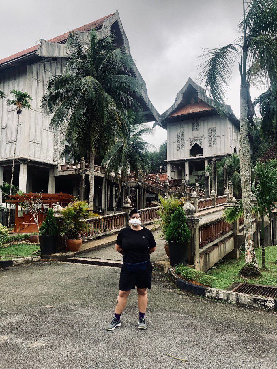 Me wearing a face mask, dressed in black tshirt and shorts, standing in front of the Terengganu state museum ramp. The building is made of several tall white traditional Malay archicture.